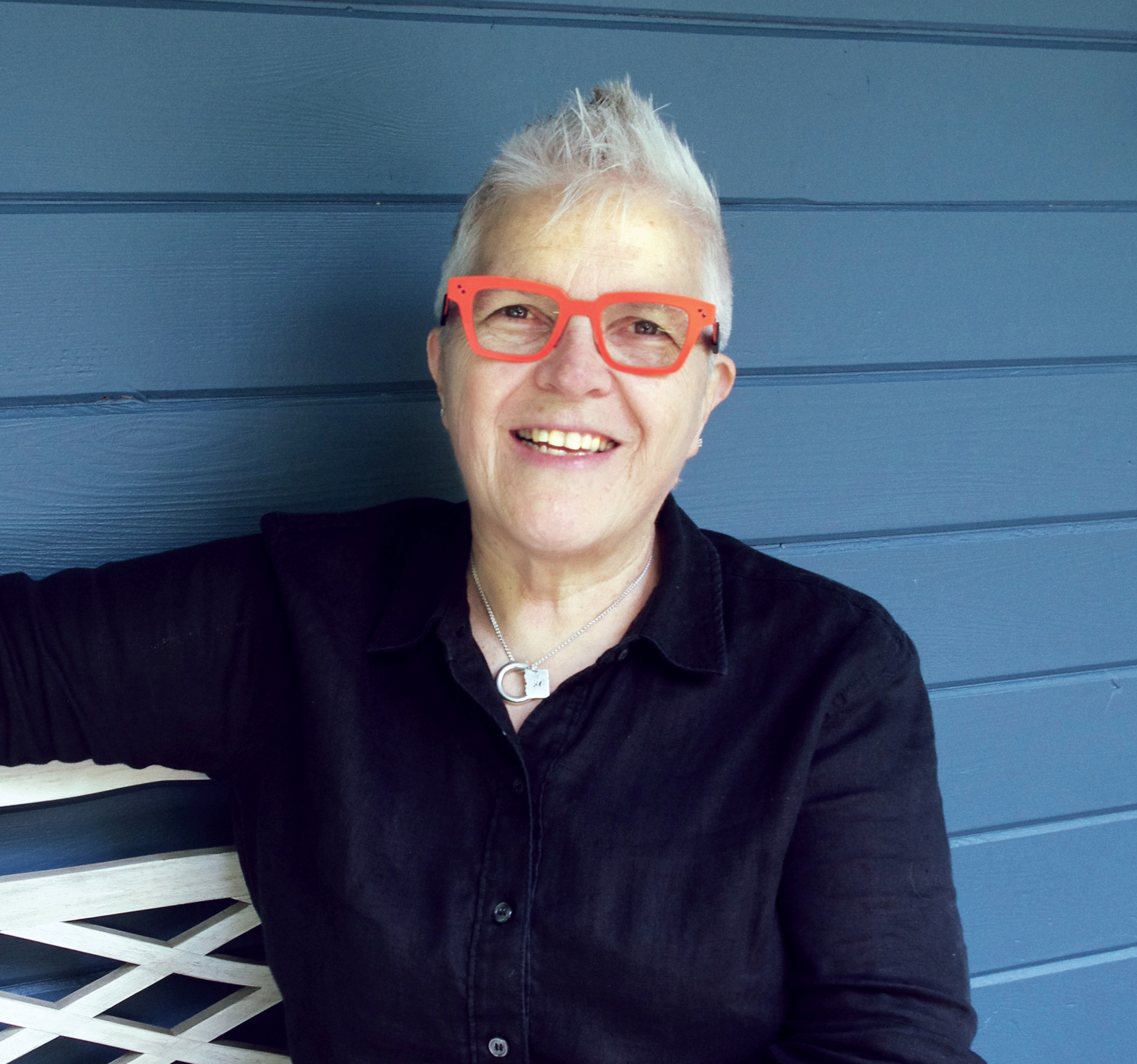 Val sitting, with dark shirt, orange framed glasses, in front of blue background